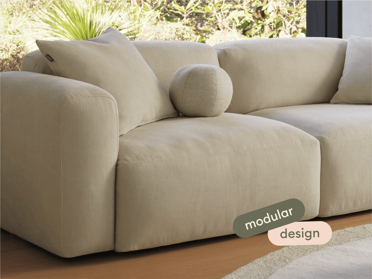 Luxurious modular sofa in beige linen fabric with plush cushions and rounded armrests, set in a sunlit modern living room with hardwood flooring.