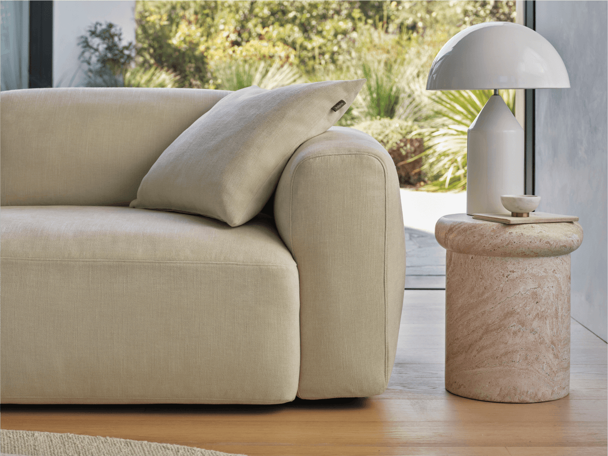 Luxurious beige linen sofa with plush cushions, clean lines, styled with a travertine side table. Elegantly placed in a sunlit living room.