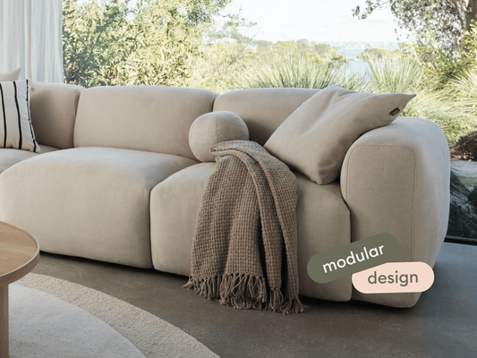Cream modular fabric sofa with plush cushioning and rounded armrests. Draped with a textured taupe throw, set in a bright, modern living room.