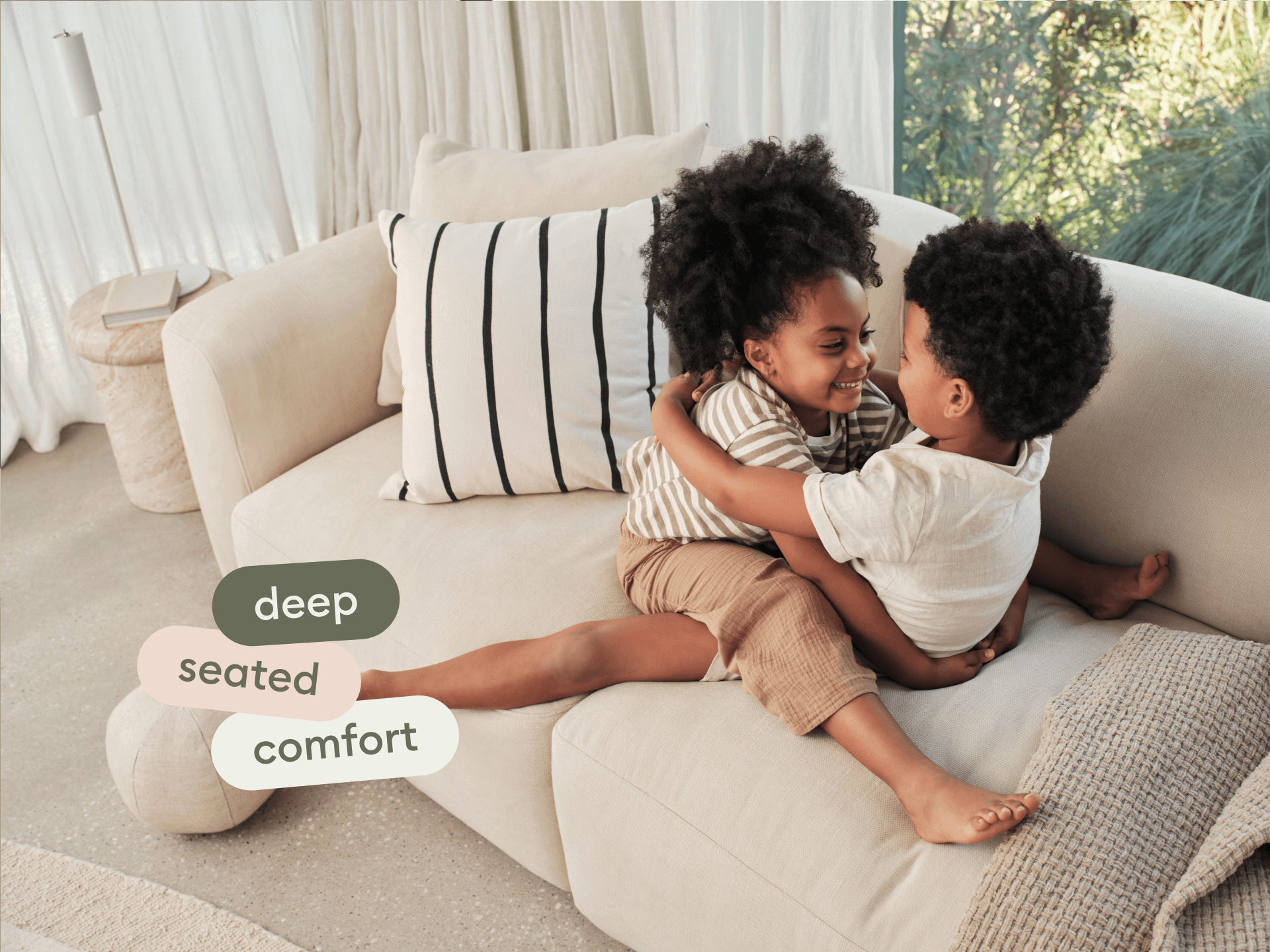 Luxurious beige fabric sofa with deep seating, plush cushions, and modern design, set in a bright, airy living room with sheer curtains and stone flooring.
