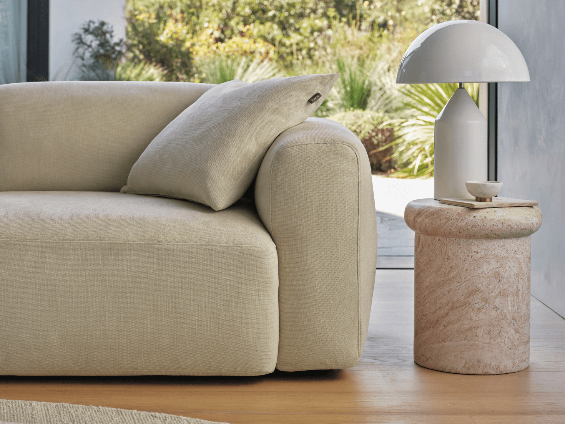 Beige linen sofa with plush cushions, paired with a round travertine side table. Modern setting features natural light and lush greenery outside.