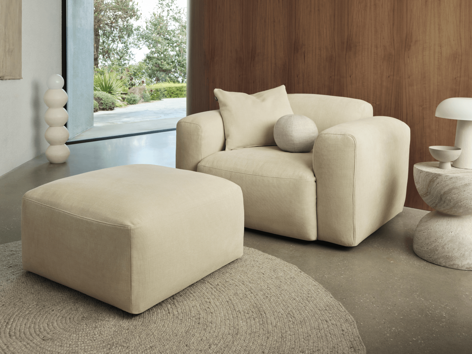 Beige modern armchair and ottoman in plush fabric, featuring clean lines and cushioned aesthetics. Set on a textured rug within a minimalist room.
