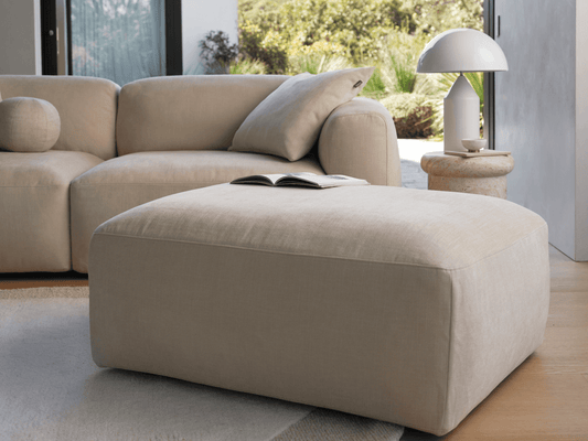 Elegant beige fabric ottoman with soft rounded edges, seamlessly blending with a plush sofa in a sunlit room featuring natural wood flooring and modern decor.