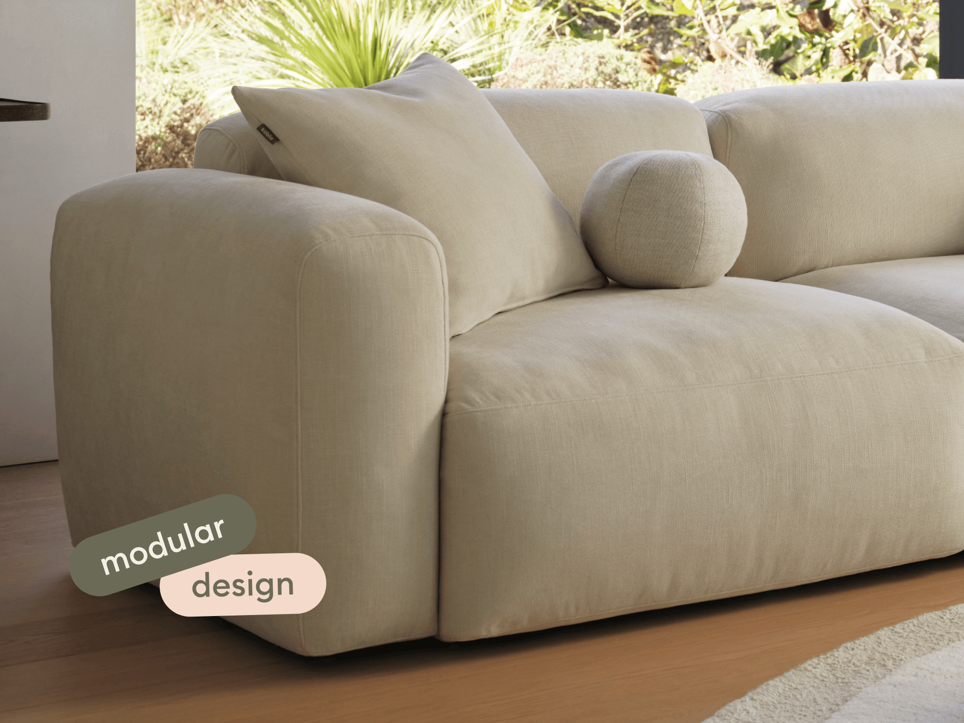 Beige linen modular sofa with rounded edges, plush cushions, and a minimalist design. Set in a sunlit living room with hardwood floors and garden view.