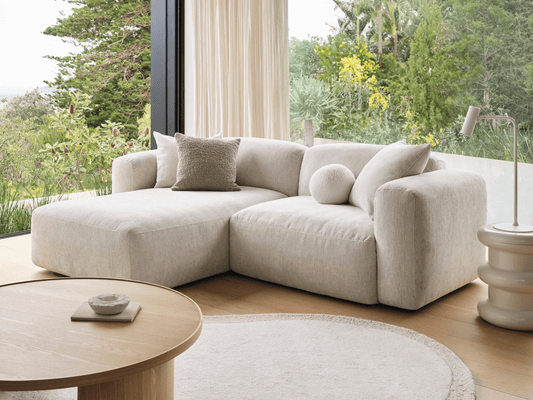 Luxurious cream linen sectional sofa with plush cushions, elegantly set in a light-filled living area featuring wooden flooring and large windows showcasing lush greenery.