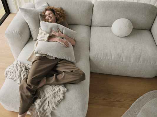 Luxurious beige sectional sofa with plush upholstery and sleek lines, set on light timber flooring. Accompanied by textured cushions and throw.