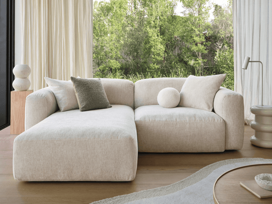 Luxurious L-shaped sectional sofa in soft beige upholstery, featuring plush cushions. Set in a serene room with timber flooring and lush greenery view.