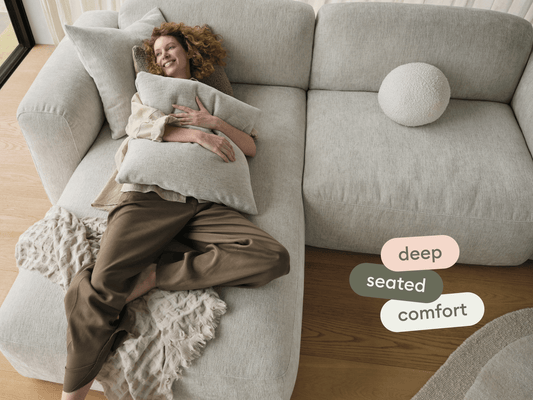 Luxurious L-shaped sofa in light grey fabric, featuring deep seating and plush cushioning. Set in a modern, sunlit living room with wood flooring.