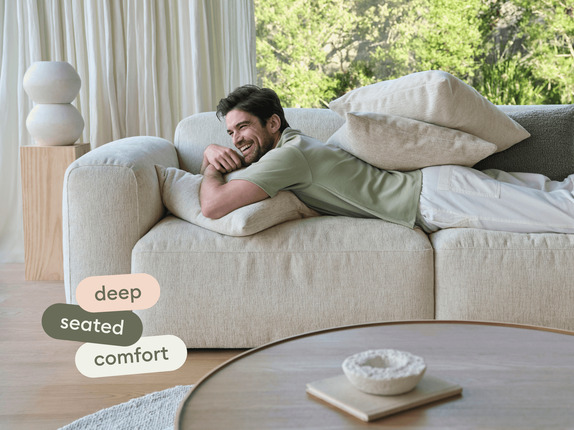 Luxurious beige linen sofa with deep seating, plush cushions, and sleek lines. Set against sheer curtains, complemented by wooden accents in a serene living space.