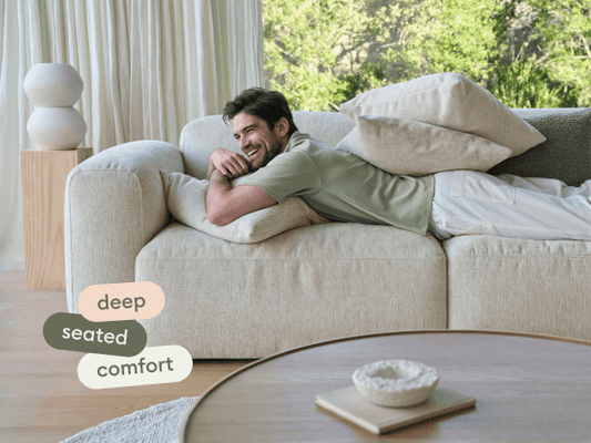 Luxurious cream fabric sofa with deep seats and plush cushions, set in a sunlit living area with light wood accents and floor-to-ceiling curtains.