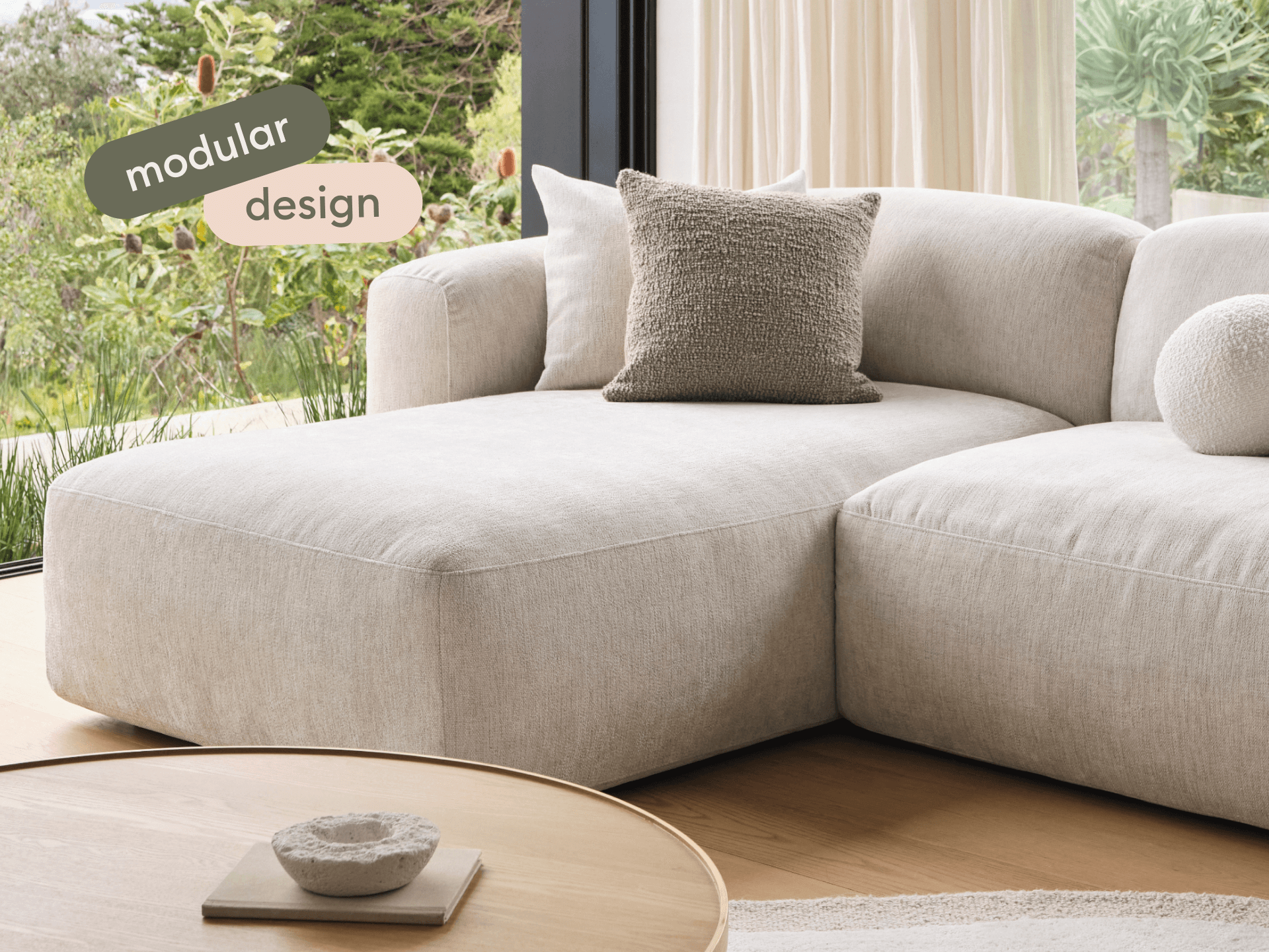 Luxurious modular sofa in soft beige fabric, featuring plush cushions. Positioned near large windows with natural views, complemented by a round wooden coffee table.