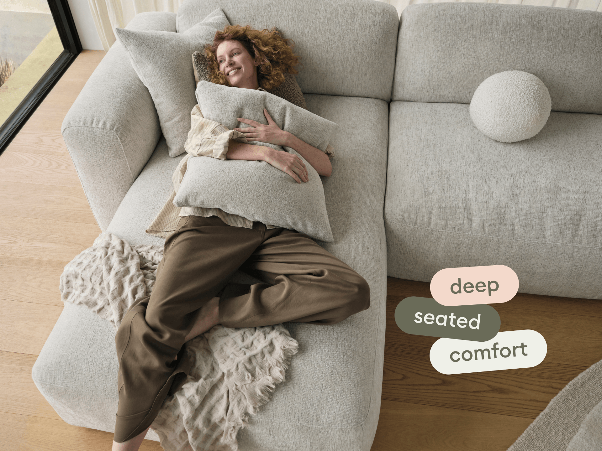Luxurious sectional sofa in soft grey fabric, featuring deep seating and plush cushions. Set on warm timber floors with natural light from large windows.