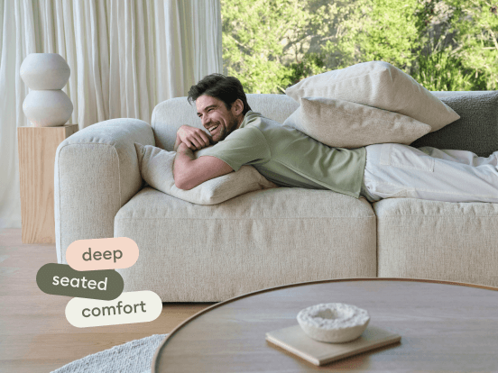Luxurious beige fabric sofa with deep seating and plush cushions; set in a bright living room with sheer curtains and a round wooden coffee table.