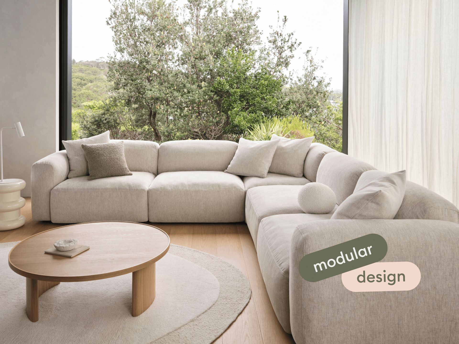 Modular sectional sofa in textured beige fabric, featuring plush cushions and curved armrests; set in a sunlit room with a round wooden coffee table.