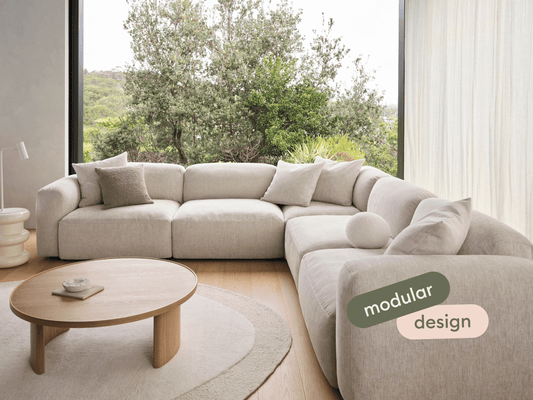 Modular sectional sofa in textured beige fabric, featuring plush cushions and curved armrests; set in a sunlit room with a round wooden coffee table.