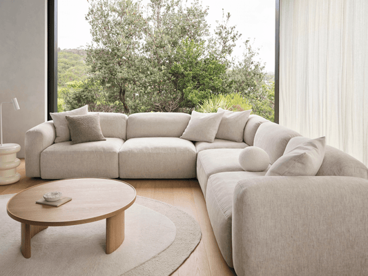 Modern L-shaped sectional sofa in soft beige fabric, featuring deep seats and plush cushions. Set in a bright room with light wooden flooring and views of greenery.