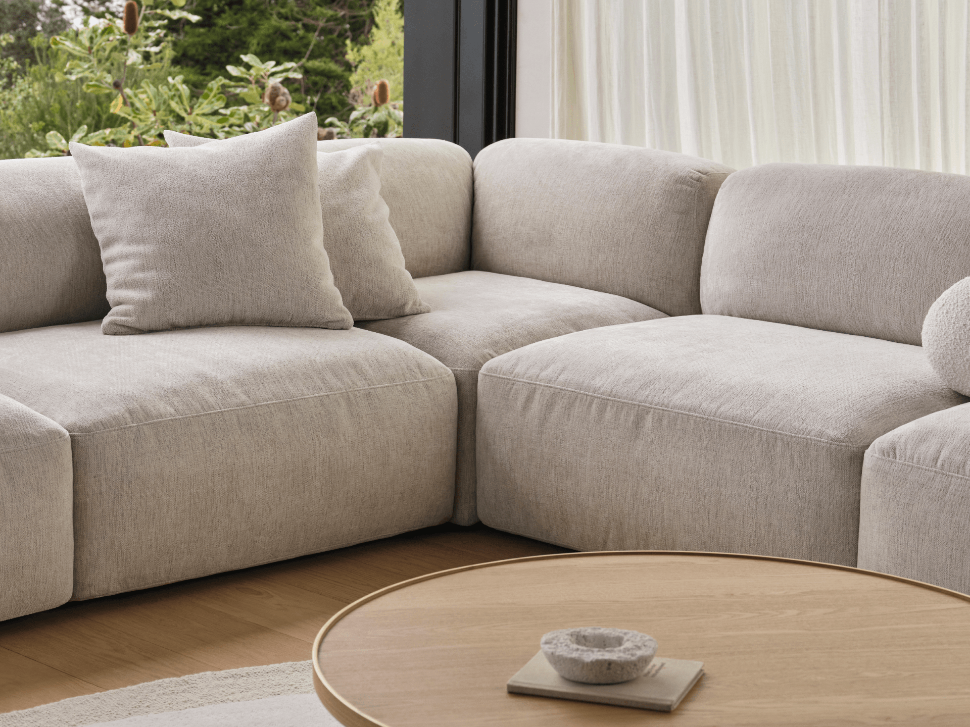 Luxurious light grey sectional sofa in soft linen fabric, featuring deep seating and plush cushions. Set against large windows, complemented by a round oak coffee table.