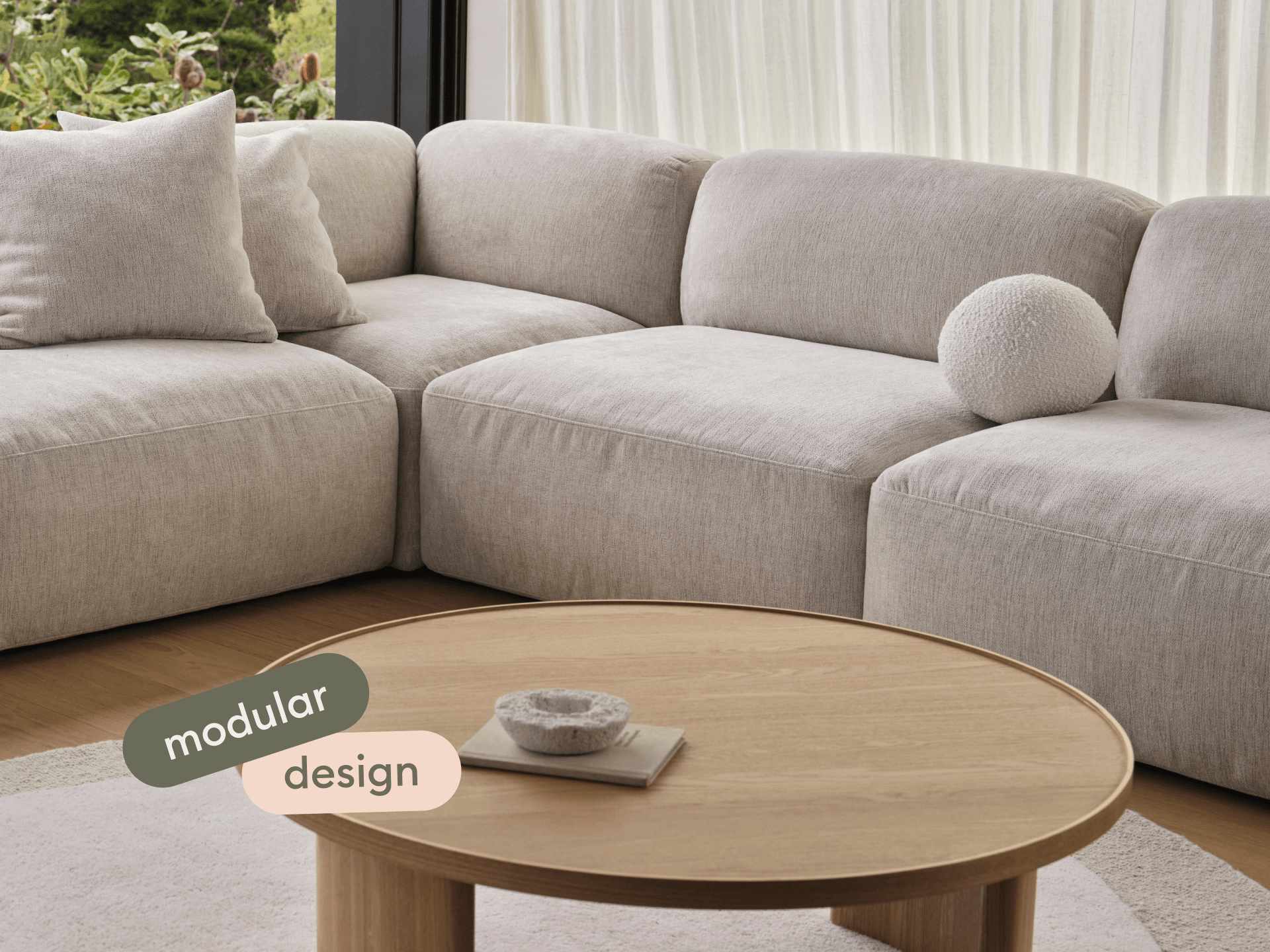 Elegant modular sectional sofa with plush beige upholstery, complemented by a round oak coffee table. Set in a bright, airy living room with serene views.