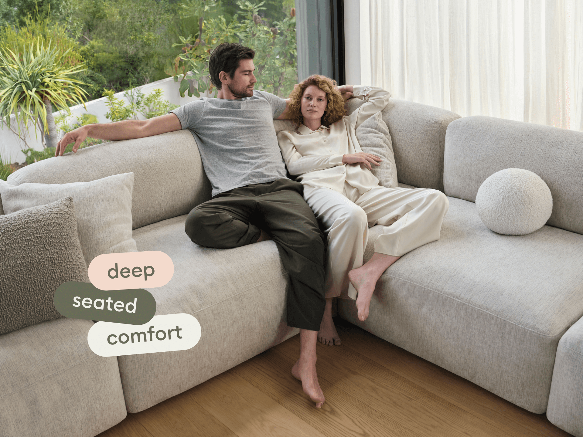 Modern beige sectional sofa with deep seating, plush upholstery, and smooth angles. Positioned on wooden flooring, surrounded by natural light and greenery.