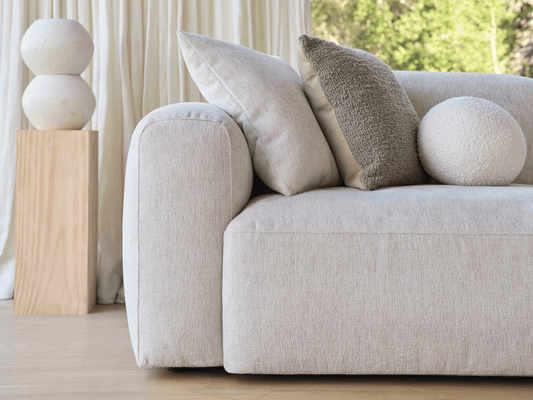 Luxurious beige linen sofa with rounded arms and plush cushions, accented by a wooden pedestal in a sunlit living area. Curtains and greenery outside.