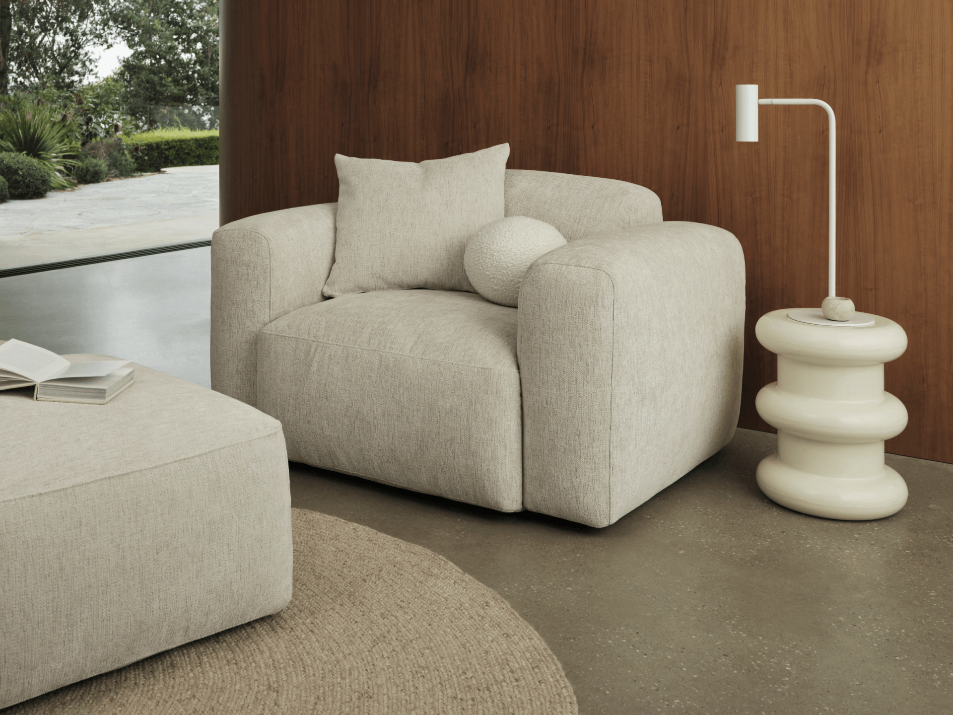 Luxurious beige fabric armchair with plush cushions, paired with a sculptural white side table. Set against a stylish wood-panelled wall and concrete floor.