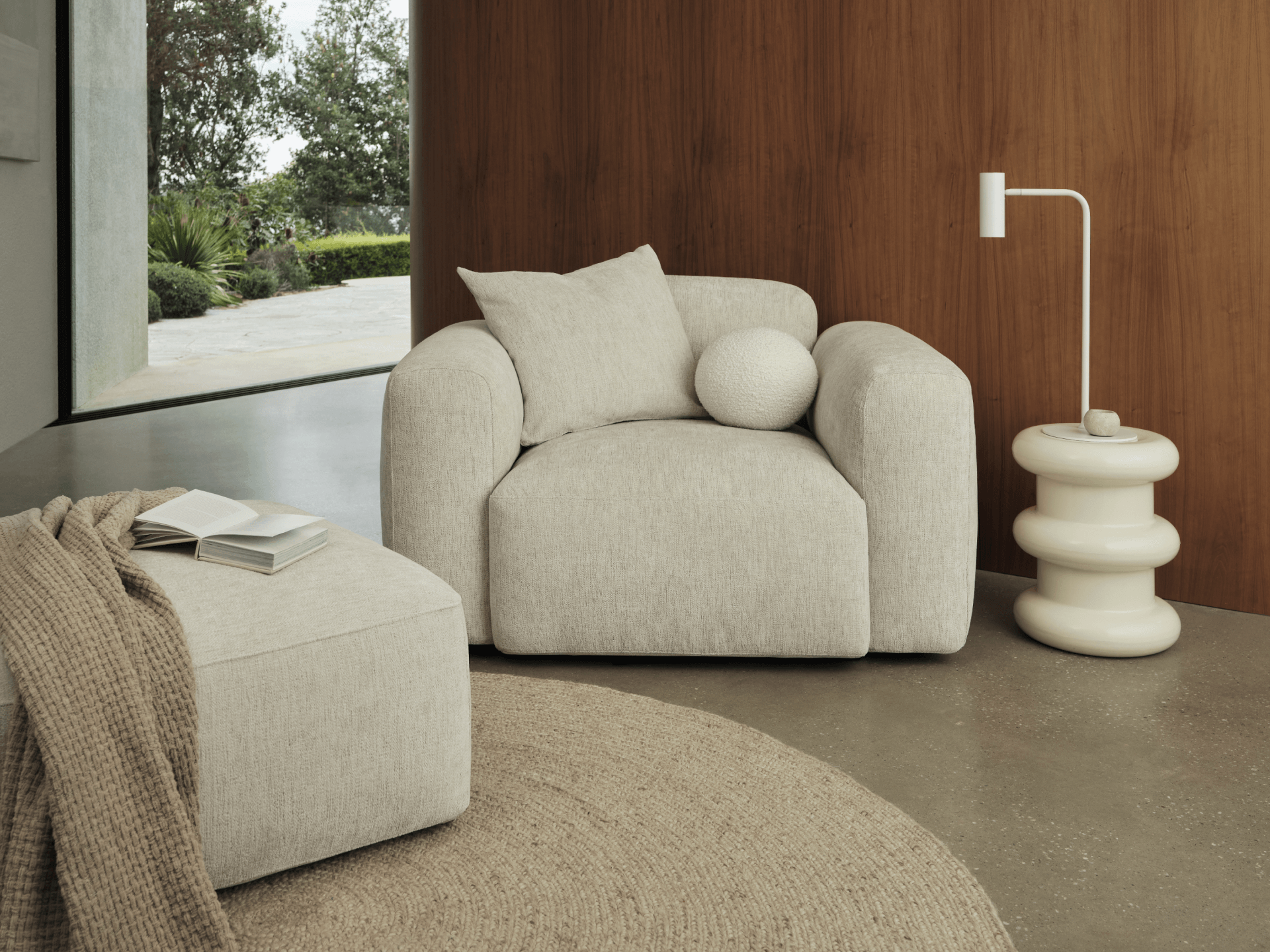 Luxurious beige armchair with plush cushions in soft fabric, paired with a matching ottoman. Set on polished concrete, accented by warm wood panelling.