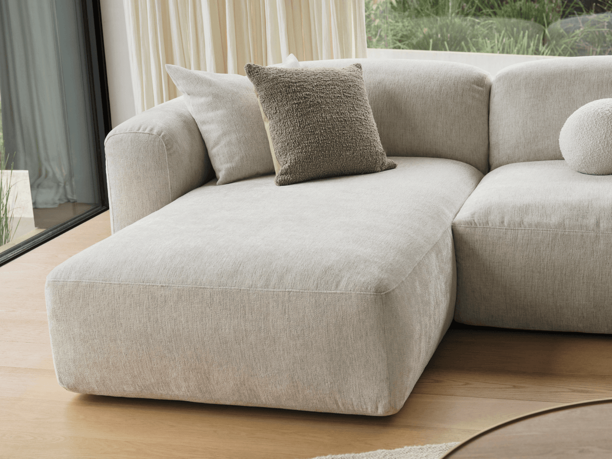 Luxurious beige fabric chaise lounge with plush cushions, elegantly set in a sunlit room with hardwood floors and soft curtains, offering a serene ambience.