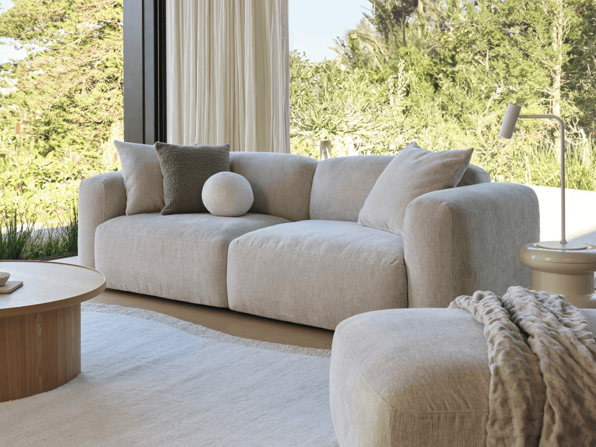 Plush beige linen sofa with curved arms, accompanied by a round wooden coffee table. Set in a sunlit room with large windows and a nature view.