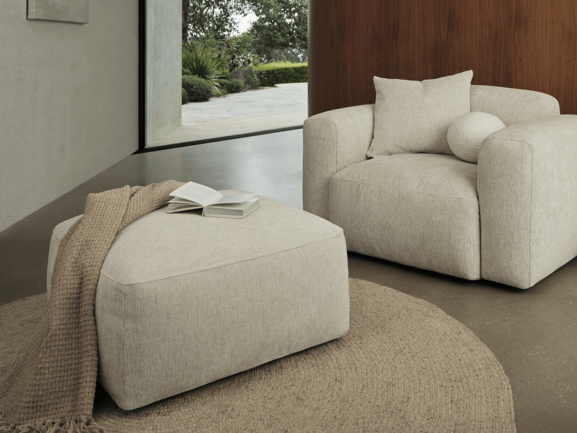 Beige fabric armchair with plush cushions and matching ottoman, set on a textured round jute rug in a contemporary living room with a wooden accent wall.
