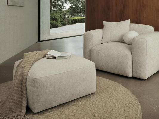 Beige fabric armchair with plush cushions and matching ottoman, set on a textured round jute rug in a contemporary living room with a wooden accent wall.