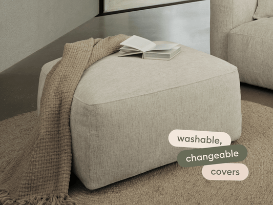 Beige fabric ottoman with washable, changeable covers. Square design, refined seams. Set on a natural fibre rug, styled with a textured throw.