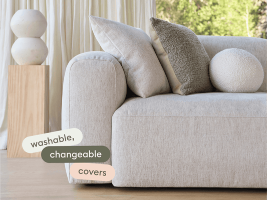 Modern beige fabric sofa with plush cushions, featuring washable and changeable covers. Set in a bright, airy room with wooden accents.