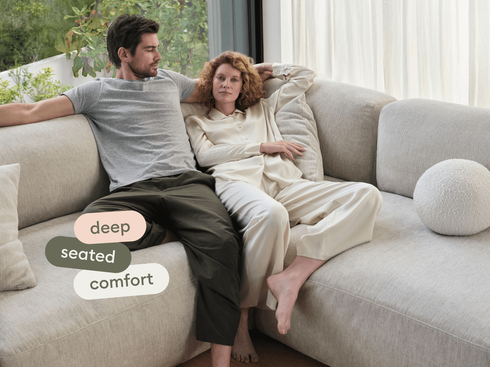 Modern sectional sofa in soft cream fabric, featuring deep seats and plush cushioning. Positioned in a cosy living room with large windows and greenery.