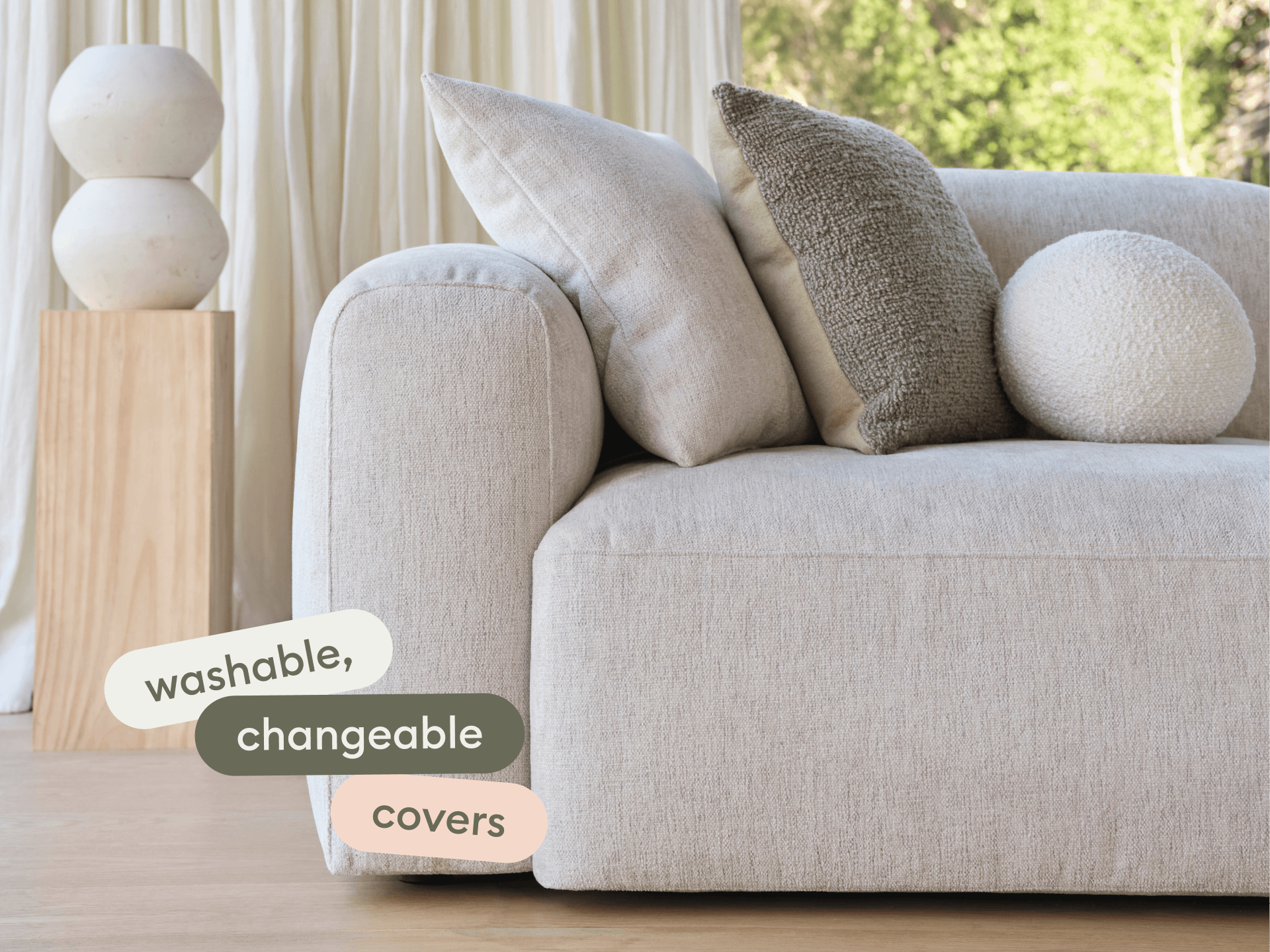 Modern beige linen sofa with plush armrests, set in a sunlit room. Features washable, changeable covers. Accented by textured cushions and wooden decor.