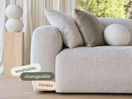 Modern beige linen sofa with plush armrests, set in a sunlit room. Features washable, changeable covers. Accented by textured cushions and wooden decor.