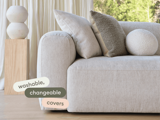 Modern beige fabric sofa with plush, washable covers. The setting features neutral cushions and a wooden pedestal with ceramic decor, in a bright, airy room.