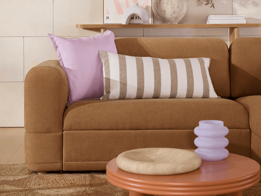 Modern brown fabric sofa with plush cushions in lavender and striped patterns. Set against neutral tones, complemented by a round wooden coffee table.
