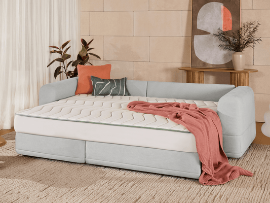 Luxurious light grey sofa bed with plush upholstery, featuring a sleek quilted mattress. Set in a chic living room with stone walls and earthy accents.