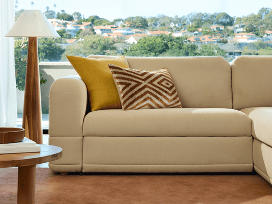 Luxurious beige modular sofa with plush fabric upholstery, curved armrests, and contrasting geometric cushions; set in an elegant living room with a scenic view.
