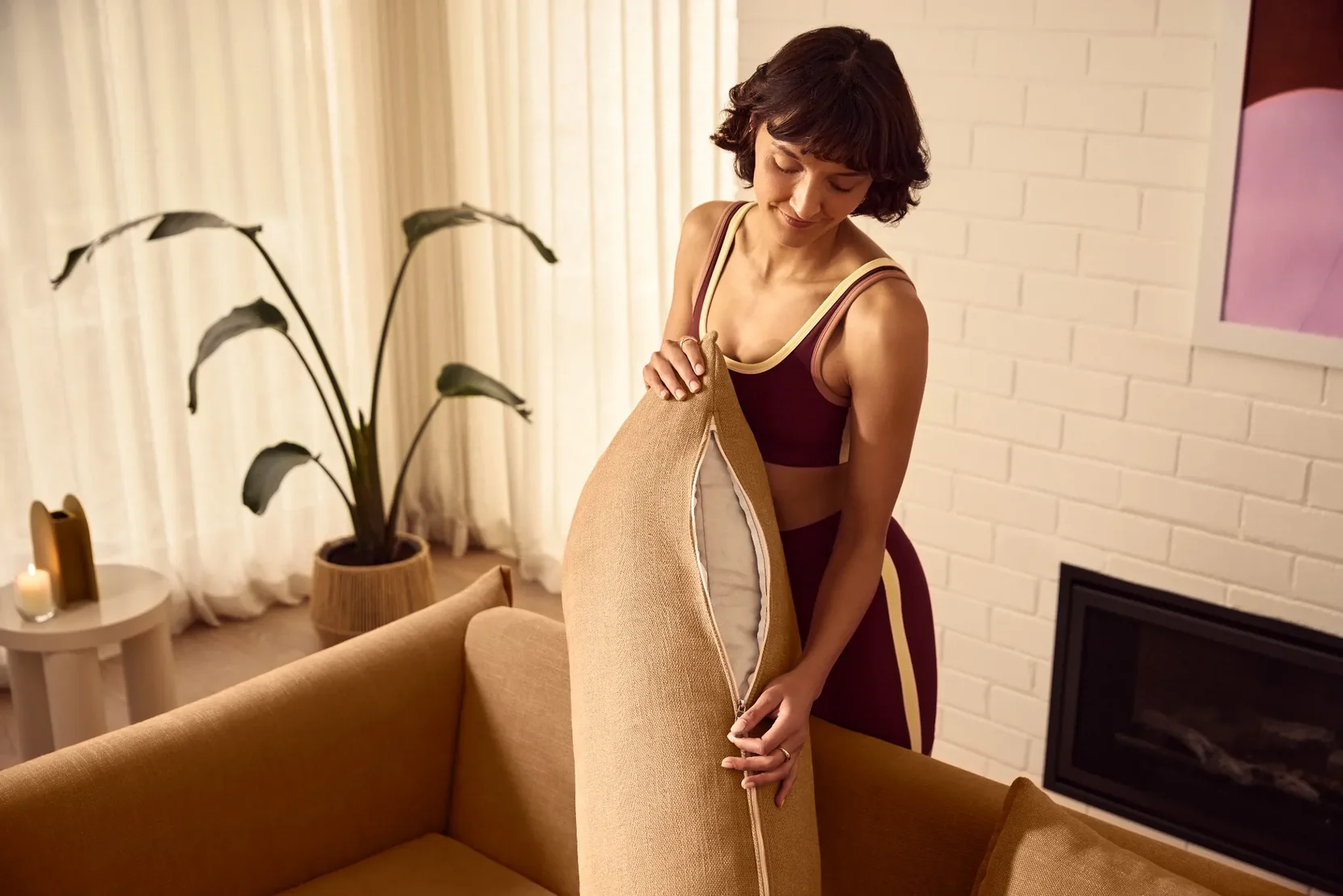 Woman holding a beige pillow in a cozy living room with a fireplace and plants.