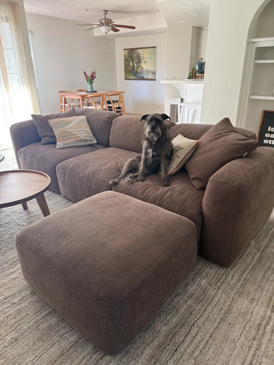 Luxurious brown fabric sectional sofa with plush cushions and matching ottoman, set on a textured neutral rug in a stylish open-plan living area.